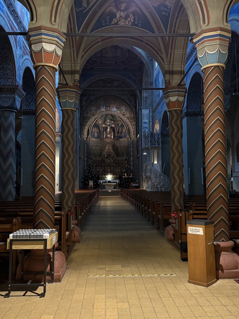 View from back of St. Mary's Church in Gengenbach, Germany. Textured columns in brown and gold support a tall archway on a center aisle between rows of pews.