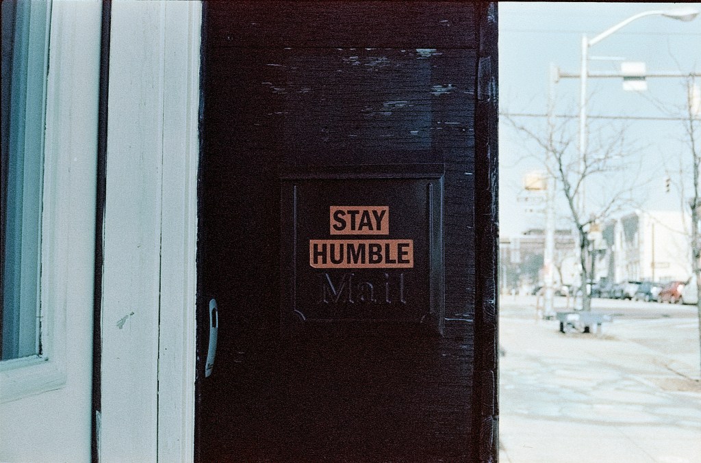 dark wood door with sign in black letters and orange background, "stay humble"